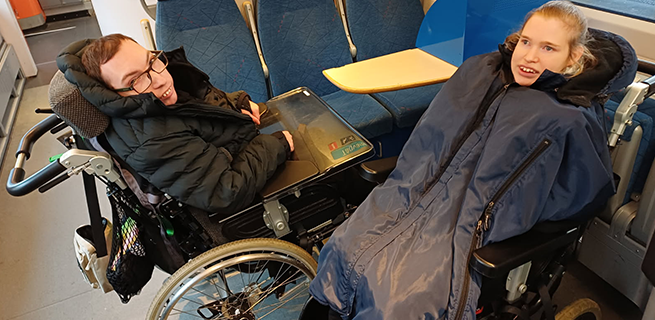 Två personer sitter i varsin rullstol bredvid varandra på ett tåg