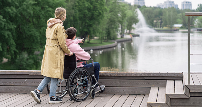 En tjej står på en brygga. Framför henne sitter en kille i rullstol och pekar ut mot vattnet.