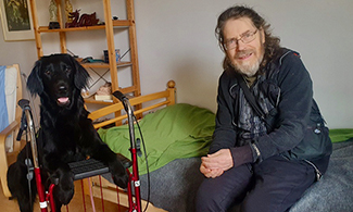 Ulf Ärnström med vårdhunden Malte. Malte står med tassarna på en rullator och Ulf sitter på en säng.